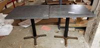 Two gray square metal-top tables side by side with black metal pedestal bases, situated in a storage or basement setting.