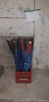 Box containing black and red metal shelf pieces wrapped in blue plastic, placed on concrete floor against wall.