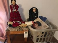 Photo showing dolls placed on and near a wooden stool with a patterned fabric seat, along with a white laundry basket containing miscellaneous items.