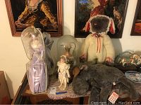 Three Franklin Heirloom dolls displayed on table including one inside plastic protective cover, with porcelain vase inside glass tube, and two stuffed animals (white and gray dogs)