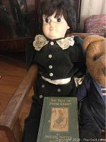 Doll with dark hair wearing a black velvet dress with lace collar sitting on a wooden bench with a small 'Peter Rabbit' book displayed in front