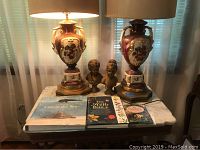 Two porcelain floral lamps with brass bases and two brass busts of children on a table, along with three books lying flat.