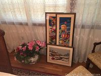 Three framed needlepoint artworks leaning against wall near window with lace curtains, alongside ceramic vase with pink faux flowers.