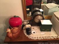 Photo showing the apple-shaped and hen-shaped cookie jars, mantle clock, and other small items on a wooden dresser with a lace doily beneath.