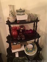 Shelf containing various glassware items including red and purple pitchers and bowls, tall white floral vase, and other decorative glass pieces