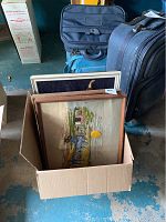 Photo showing 4 framed vintage wall art pieces packed standing inside a cardboard box on a floor with suitcases and bags in background.