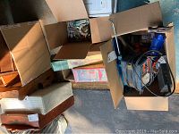 Boxes containing various hand tools, garden supplies, bird feeders, and wind chimes seen outdoors on pavement.