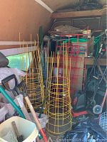 Photo showing several tall yellow wire tomato cages clustered together beside some red wire square-frame tomato cages with a shovel and other gardening tools nearby.