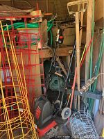 Photo of black and red Yard Machine rototiller in storage with gardening tools and wire fencing around it.