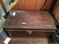 Front and top view of a rectangular wood chest with intricate hand carved scenes on top and sides, showing detailed figures and botanical elements. The chest has a dark finish and a brass latch.