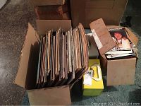 Two cardboard boxes containing approximately 60 LP vinyl records and over 100 45 RPM vinyl records in varying condition, standing vertically with album covers visible.