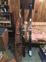 Photo shows a pair of vintage wooden snowshoes with rawhide webbing and leather bindings, Bear Archery compound bow in black, and a pair of black alpine skis with bindings leaning against a couch.