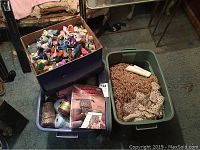 Photo of three boxes containing crafting materials: one with needlepoint thread, one with assorted yarns including macrame cord and crochet yarns, and another with folded table cloths and lace fabrics, plus a small woven pet home.