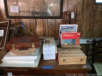 Overview of painting supplies lot showing books, easel boards, and storage boxes including a brown suitcase-style box with floral design and a large wooden box labeled Morton Chest.
