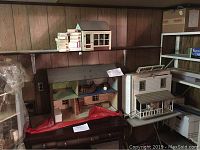 Three doll houses arranged on shelves and a dresser. One is smaller with a blue roof and visible interior furniture, another is larger with a green roof, and the third is white with a porch and figures inside.