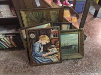 Five framed pieces of wall art leaning against a step, shows various artwork including embroidery of girl reading, ship painting, and other framed paintings