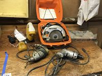 Four power tools on wooden surface including Skilsaw circular saw and orange case, yellow hand sander, and two electric drills