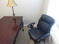Overall front-side view of black upholstered office chair next to wooden desk and lamp.