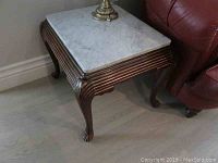 Pair of end tables with carved dark wood bases and light Carrera marble tops, positioned between pieces of leather furniture, showing overall condition and details of tops and bases.