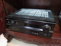 Front and top view of the black Yamaha receiver showing control knobs, display screen, speaker terminals and ventilation grilles.