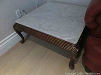 Square coffee table with white Carrera marble top and carved wood base, shown in corner beside red chair and wall outlet, highlighting the carved legs and grooved base design.