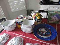 Photo showing multiple white square salad or soup bowls stacked, colorful mugs, a ceramic cookie jar with fruit basket design, and various plates including a large blue Royal Doulton Studio Touch plate.
