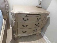 Front view of the painted wood side table showing the curved serpentine front with 3 drawers and metal drawer pulls, highlighting the ornate carved border details along the top edge and legs.