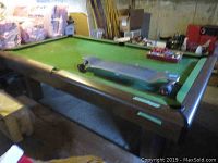 Full view of pool table with green felt, soiling visible, and some pool balls and cues on the surface and nearby.