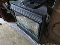 Front view of the standalone gas fireplace showing black metal frame and glass front panel with plastic covering inside.