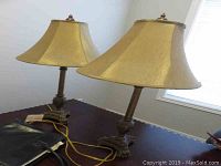 Pair of decorative table lamps with golden-beige fabric shades placed on a dark wooden surface, cords visible.