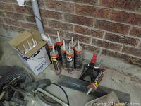 General view of construction adhesive tubes and some tools on floor against brick wall.