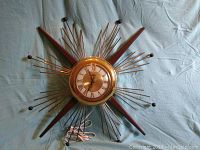 Full view of starburst wall clock showing brass rods and wooden accents, with white Roman numeral clock face and clock hands, cord visible.