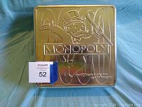 Top of rectangular metal game box with embossed Monopoly text and Mr. Monopoly character, with '2000' in background, silver metallic finish.