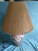 Full view of the lamp showing the textured pottery base with white and brown mottled glaze and the tall woven natural fiber conical shade on a blue background.