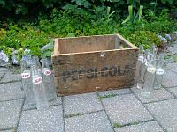 Wooden Pepsi-Cola crate with 9 assorted vintage glass bottles arranged around it on outdoor pavement.