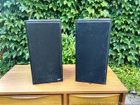 Pair of Paradigm Model 3se bookshelf speakers with black cloth grills on, placed outdoors on wooden surface with greenery background.