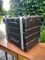 Full view of black hard carrying case on wooden table outdoors with greenery background.