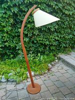 Full view of lamp outdoors on brick patio, ivy backdrop showing curved teak stand, round base and conical shade.