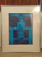 Framed abstract mixed media artwork with blue and brown geometric design inside a thin metallic frame with white mat.