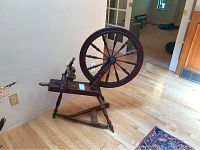 Side angle of the vintage spinning wheel showing the wheel and treadle on a wooden floor.