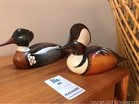 Two handcrafted wooden bird sculptures on a table, identified as Red Breasted Merganser and Hooded Merganser.