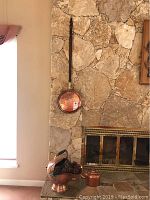 Wide view showing copper warming pan hanging on stone wall above brass fireplace screen, large copper coal bucket filled with pine cones, and copper kettle nearby.