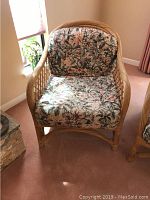 Wicker indoor chair with floral seat and back cushions against a carpeted room with window light.