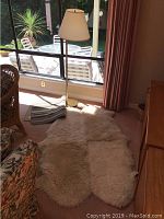 Sheepskin rug spread on carpet floor near a window, with floor lamp next to it and small striped rug partially visible