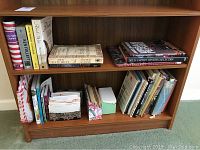 Wooden bookshelf with two shelves filled with books, some folders, and binders, showing a variety of book spines and titles.