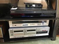 Four pieces of vintage electronics stacked on shelving: Sony Blu-ray player, Akai double cassette deck, Sansui AM/FM tuner (silver), Technics 5-disc rotary CD changer with remote on top.