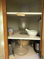 Cabinet shelf with clear glass decanter, white ceramic flour sifter, metal grater, mixing bowls and other kitchen items