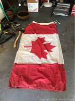 Photo of Canadian flag showing red maple leaf on white background with red side panels, laid on floor with visible damage, wrinkle and tear