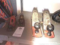 Photo showing four hand planes on a shelf, including two smaller red-based planes and two larger metal planes with black knobs and handles spaced side by side.