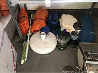 Photo showing two rolled orange tarps, a small tent with frame poles, a white round collapsible base, and additional camping supplies, including a blue bag.